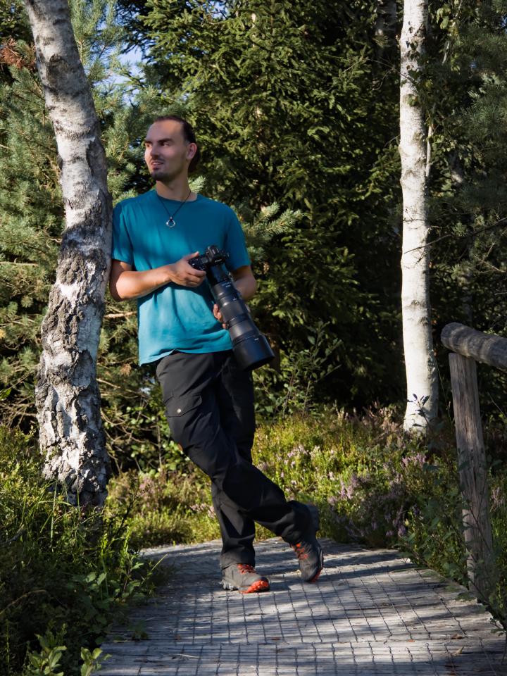 A young man (Lukas Nagel) with a short beard in a teal t-shirt and dark hiking trousers leans against a white birch trunk on a narrow wooden boardwalk, holding a Nikon camera with a long telephoto lens in both hands. He looks off to the left, lit by low afternoon sun, with dense green conifers, birches and flowering purple heather around him.