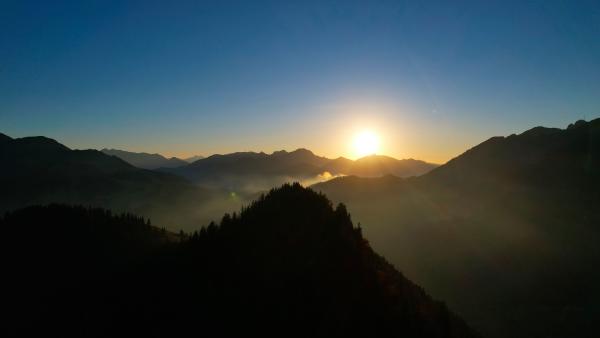 Drone photo taken over the mountains pointing at the sun which is setting among them.
