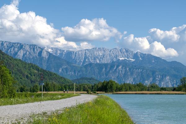 A trail with two ciclists on it to the left, a rivert to the right and some mountains in the background.