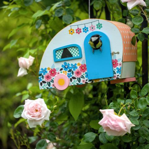 This is a photo of a cute bird with a little bird house in a caravan style. The bird house is hanging in a rose bush so the background is green with some roses.