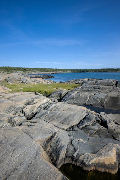 There is a rocky beach with some little ponds in it. You can see a green meadow in between the rocks. Behind it there is the sea and a land mass behind it.