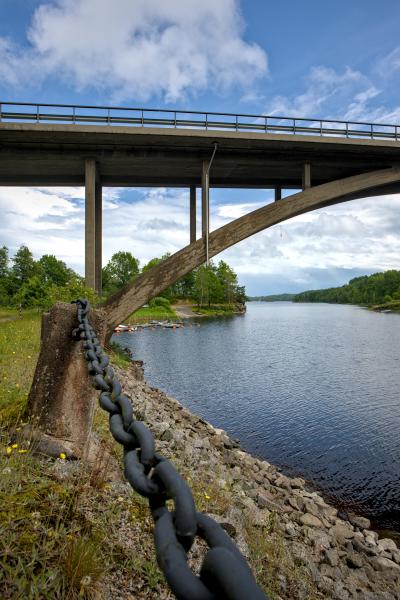 A iron chain next to a lake / river with a lot of rocks beyond it. In the background is a bridge. Around the river / lake is a forest. In the water there are some boats.