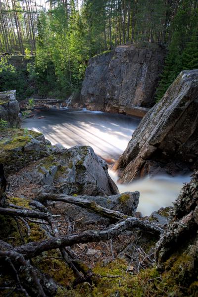A waterfall with a forest in the background. The water is washed out because of the long exposure. The sky is blue and cloudy.