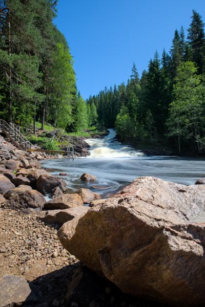 You can see a river with a waterfall. The waterfall is smooth because of long exposure. There is a forest and the blue sky. In the foreground are some rocks.
