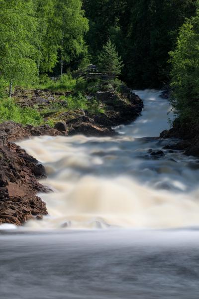 You can see a river with a waterfall. The waterfall is smooth because of long exposure. There is a forest and the blue sky. In the foreground are some rocks.