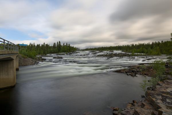 A little waterfall in a river with a bridge to the left. There is forest in the background and the sky is cloudy.
