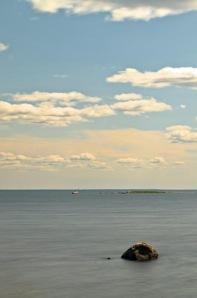 A single stone in the washed out sea with a small island in the background and a big one just at the horizon. The colors a bluish yellowish and have a soft feeling.