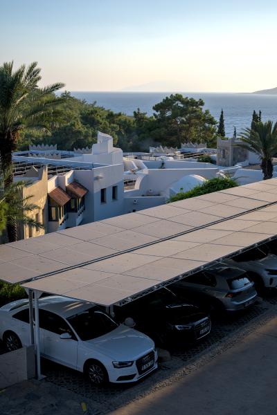 You can see a parking lot with solar panels aboth the cars. Behind that there are some white turkish buildings and in the background you can see some trees and the sea.