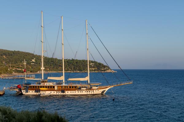A sail ship with no sails up. It is docked to land and reflecting the colors of a distant sunset.