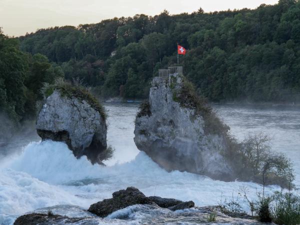 Photo of the Rheinfall in Switzerland. A Swiss flag sits on the top of a rock in the water.