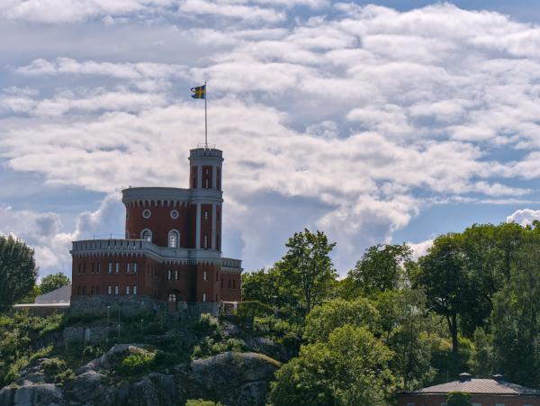 A castle on a rock with some trees around it. On top of the castle there is a swedish flag.