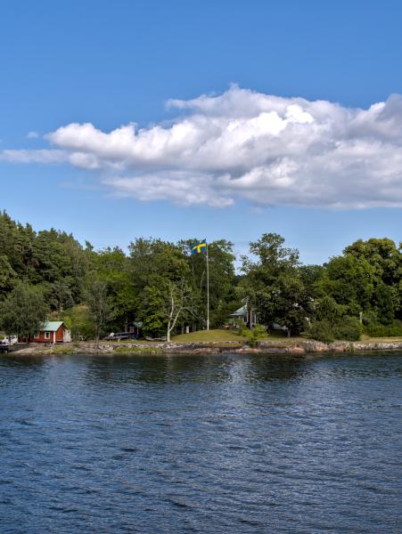 A swedish flag on a long post nex to a typical red cottage. There are some trees and the sea.