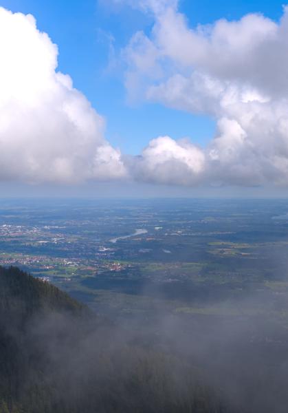 Image taken from a bavarian mountain with villages and a river below and big white clouds in the blue sky.