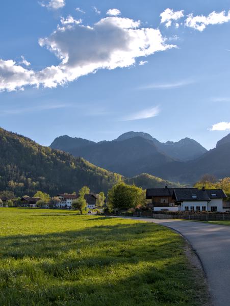 A rural scene in the bavarian mountains. There are some houses on the right of the street which snakes threw the scene. In the background are the mountains and the sky is blue with white clouds.