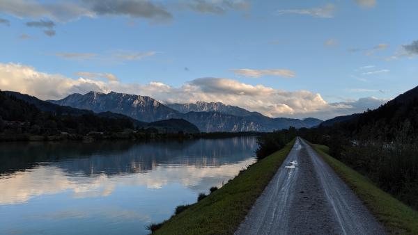 A tranquil river reflecting the sky with a path alongside it and mountains in the distance under a cloudy sky.