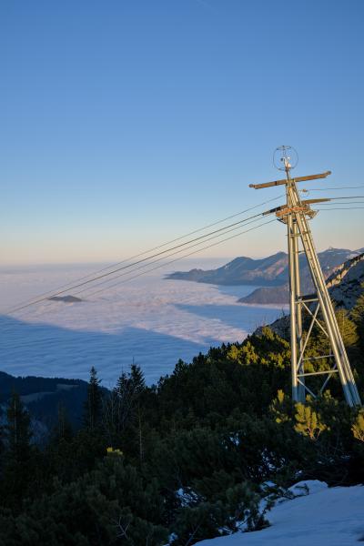 On the right side there is a pillar of a lift. Behind that there can be seen several mountains and below those mountains is a big sea of clouds till the horizon. The sky is blue with a slight orange touch.