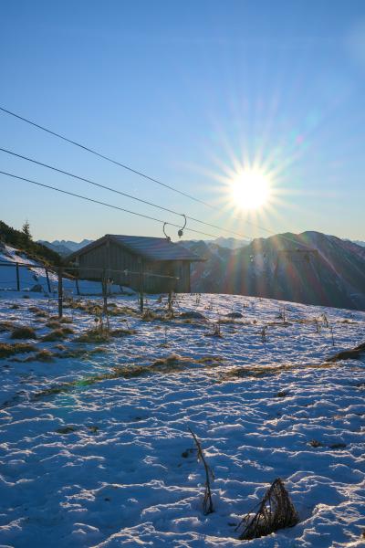 A scene taken in the bavarian mountains. In the foreground there is a sunny maedow with a mountain hut and a ski lift on it. In the background you can see the sun with a sunstar right above a mountain range. The sky is blue and a little bit orange on the horizon.