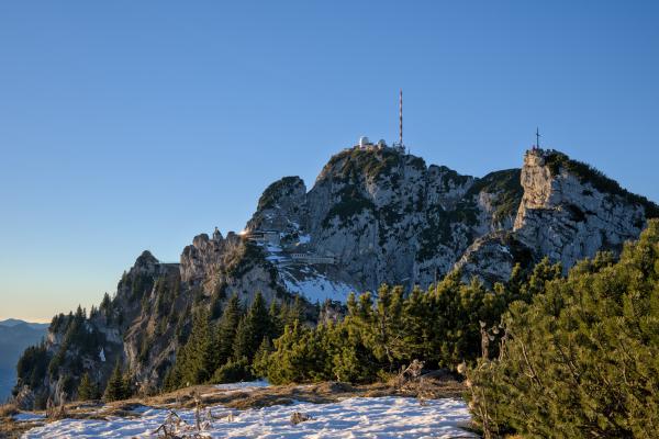 You can see the vast top of the mountain Wendelstein in the bavarian alps. In the foreground there is some snow and bushes. In the background lies the mountain top with an antenna on top of it and several buildings. On the right side of the image there is another smaller mountain top. The sky is blue to orange at the horizon.