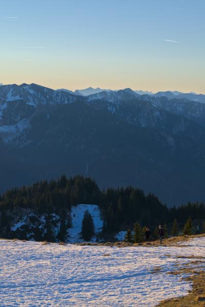 The image shows a landscape with a vast mountain range in the background. The sky has a blue / orange color. In the foreground there is a slope with snow and two small looking persons hiking.
