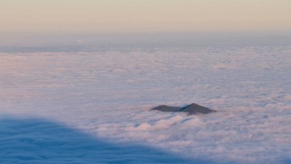 There is one mountain looking out of a infinite appearing bed of clouds. The sky is toned with a light orange.