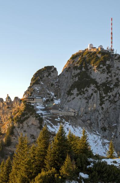 In this picture you can see the top of a high mountain with some kind of antenna on top of it. Below the top there are some buildings and at the bottom of the image there is a little forest.
