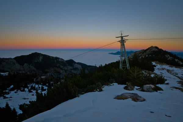 This image was taken from a mountain looking down on a sea of clouds till the horizon. Several other mountains can be seen in the background. Thr horizon is orange, purple and blue. In the foreground there is some kind of pillar of a lift.