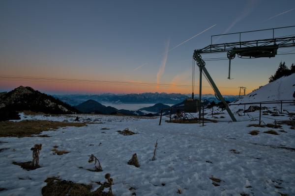 A skilift at the top of a mountain. There lays snow and in the background there are several other mountains. The sun is setting so the sky has some beautiful colors.