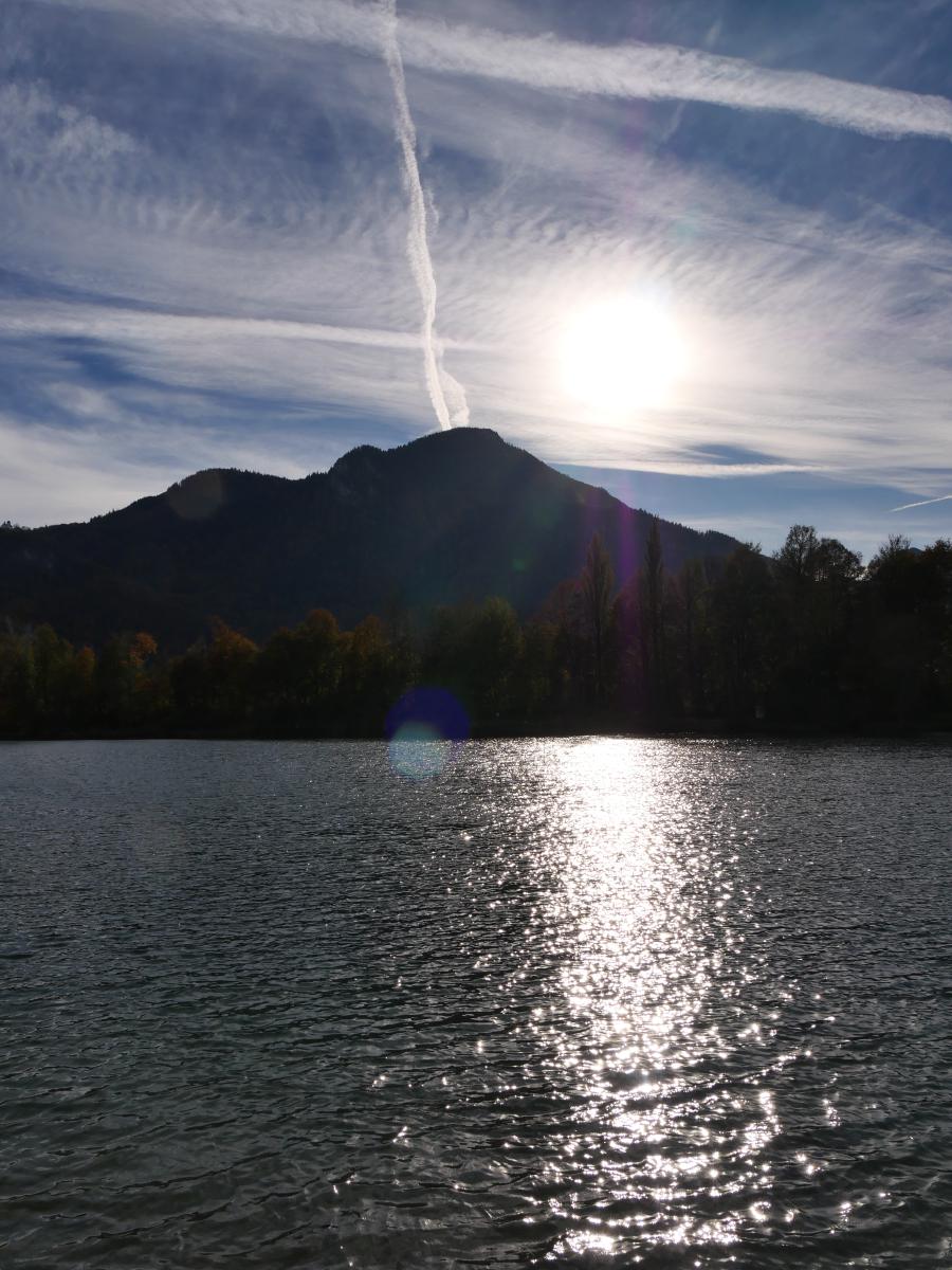 Den lågt stående solen står över ett skogklätt berg och lägger en bred glittrande stig över en sjö, medan kondensstrimmor korsar den ljusblå himlen.