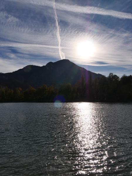Den lågt stående solen står över ett skogklätt berg och lägger en bred glittrande stig över en sjö, medan kondensstrimmor korsar den ljusblå himlen.