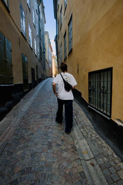 A man with a top knot, seen from behind, walks down a narrow cobbled lane in Gamla stan, Stockholm's old town. He wears a white T-shirt printed with tadpoles, a black belt bag slung across his back and dark striped track pants. Ochre-yellow houses with green shutters and a barred window rise on both sides toward a thin strip of blue sky.