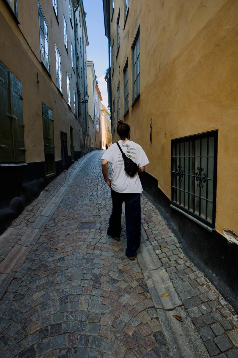 En man med hårknut, sedd bakifrån, promenerar längs en smal kullerstensgränd i Gamla stan i Stockholm. Han bär en vit t-shirt med tryck av grodyngel, en svart magväska snett över ryggen och mörka randiga träningsbyxor. På båda sidor reser sig ockragula hus med gröna fönsterluckor och ett gallerförsett fönster mot en tunn strimma blå himmel.