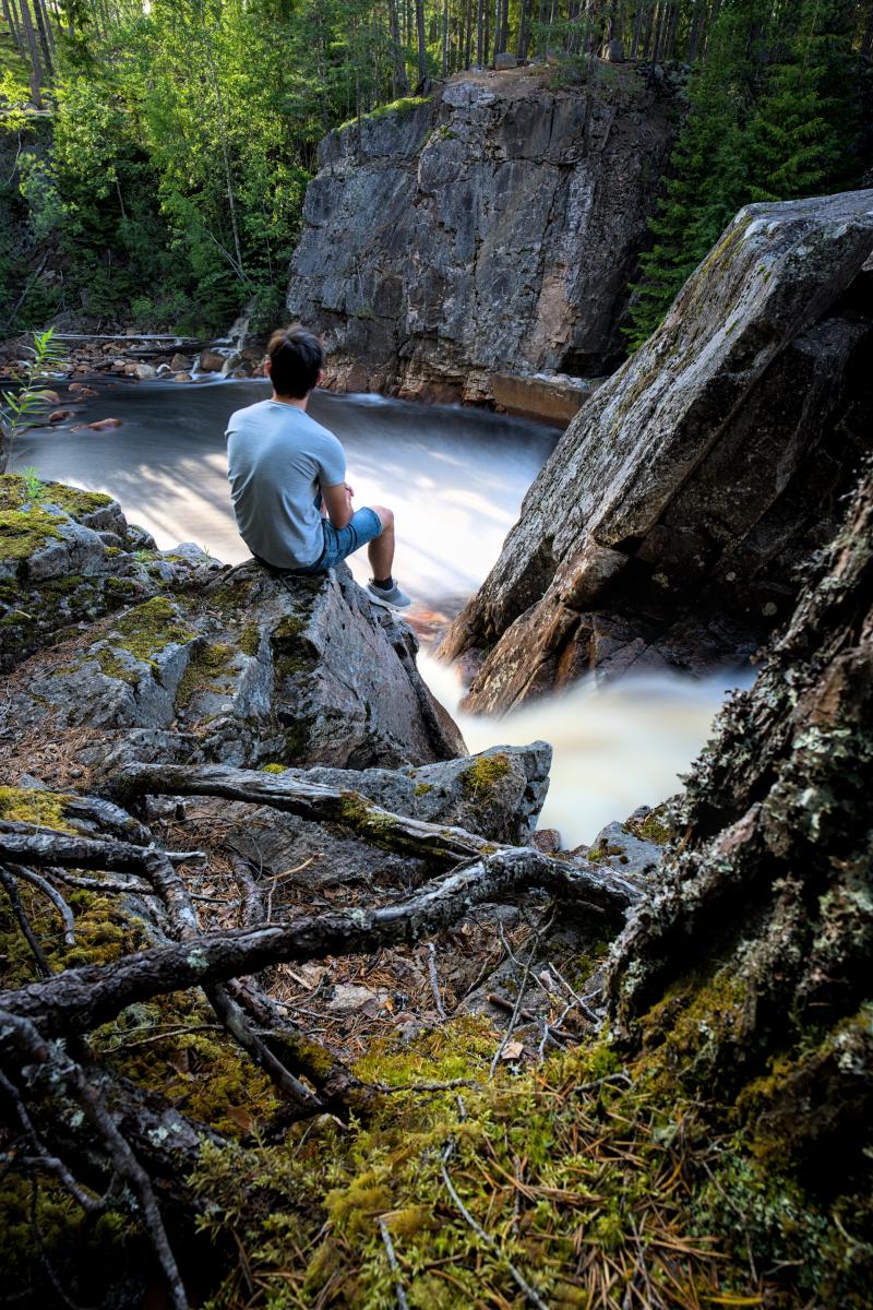 Ein Selbstporträt von mir, wie ich auf einem Felsen neben einem Wasserfall sitze. Der Wasserfall ist wegen der Langzeitbelichtung verwaschen.