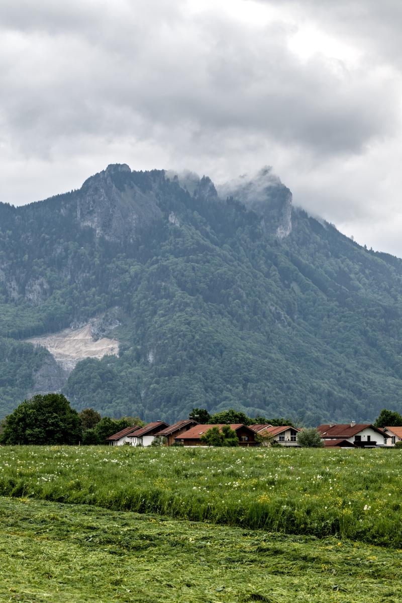 Ein Regentag in einem bayerischen Dorf mit einigen Häusern und einer frisch gemähten Wiese. Im Hintergrund liegen Berge in den Wolken.