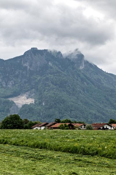 Ein Regentag in einem bayerischen Dorf mit einigen Häusern und einer frisch gemähten Wiese. Im Hintergrund liegen Berge in den Wolken.