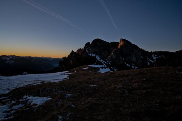 Der weite Gipfel des Wendelsteins vor einem Sonnenuntergangshimmel. Im Vordergrund liegt eine teilweise schneebedeckte Wiese.