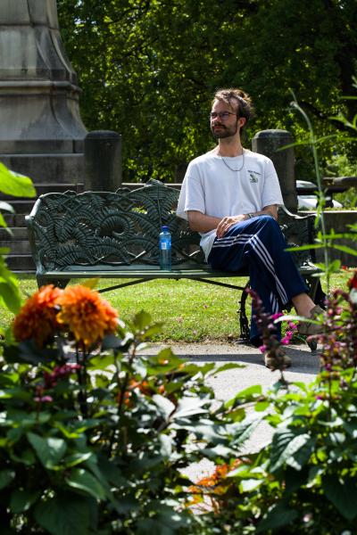 Ein bärtiger Mann mit Brille und Dutt sitzt auf einer verzierten grünen Parkbank aus Gusseisen in Stockholm und blickt in der Sommersonne zur Seite. Er trägt ein weißes T-Shirt und eine dunkelblaue Jogginghose mit Streifen. Orangefarbene Dahlien und grüne Pflanzen füllen den unscharfen Vordergrund, im Hintergrund das Laub hoher Bäume.