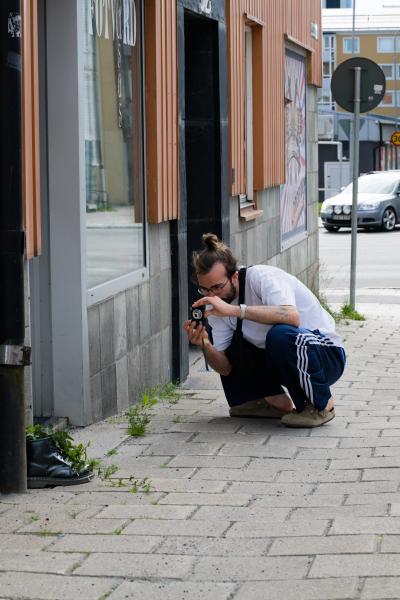 En man i vit t-shirt, träningsbyxor och hårknut sitter på huk på trottoaren i den nordsvenska staden Luleå och fotograferar ett par kängor fulla av murgröna med en liten kompaktkamera.