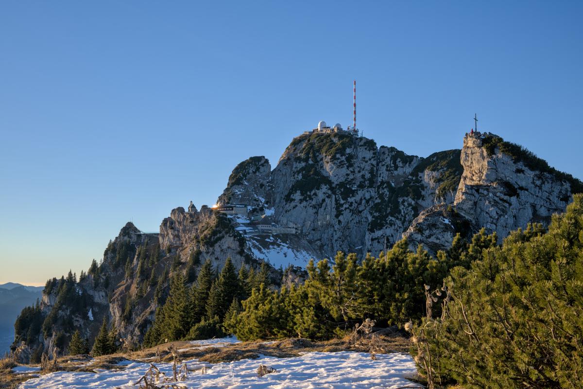 Zu sehen ist der weite Gipfel des Wendelsteins in den bayerischen Alpen. Im Vordergrund liegen etwas Schnee und Büsche. Im Hintergrund liegt der Berggipfel mit einer Antenne darauf und mehreren Gebäuden. Rechts im Bild ist ein weiterer, kleinerer Gipfel. Der Himmel ist blau und am Horizont orange.