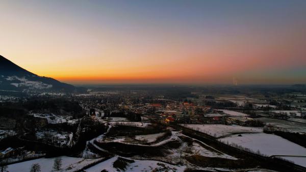 En flygvy i gryningen över en snötäckt dal: en stad med upplysta gator i mellangrunden, en mörk bergssida till vänster och ett brett orange ljusband längs horisonten.