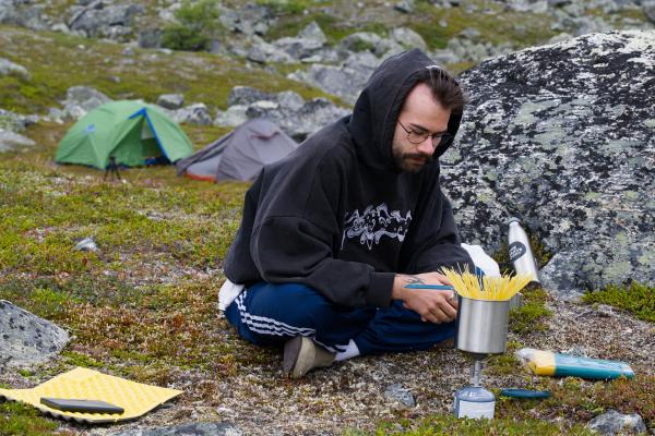 En man med glasögon och skägg, klädd i huvtröja och träningsbyxor, sitter framför ett campingkök med en kastrull full av spaghetti. I bakgrunden syns två tält som smälter fint in i det steniga fjällandskapet.