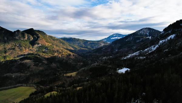 En flygvy längs en dal mellan mörka skogklädda ryggar, med kala vintersluttningar, snöfläckar och en avlägsen snötäckt topp under en gråblå himmel.