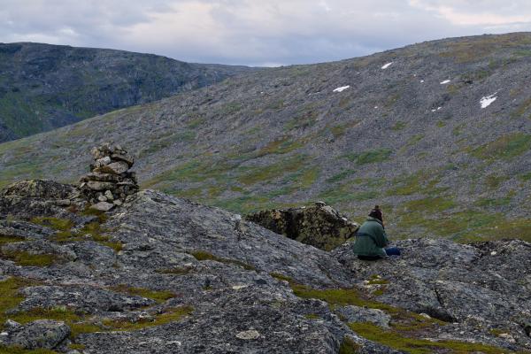 En man i jacka, träningsbyxor och hårknut sitter ensam och röker några meter från ett röse i svenska fjällen. Landskapet består av tre steniga nivåer av berg och en molnig himmel. Det känns dystert, eftertänksamt och lite deprimerande.