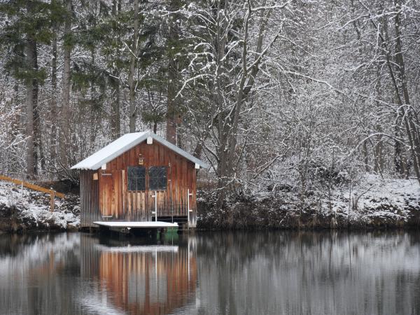 Eine schneebedeckte Hütte, die sich in einem stillen See spiegelt, umgeben von schneebestäubten Bäumen.