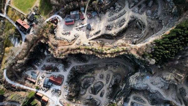 En lodrät flygvy över ett stenbrott: ljus skuren berggrund, staplar av hällar, slingrande grusvägar och några verkstadsbyggnader med röda tak, med skog i kanterna.