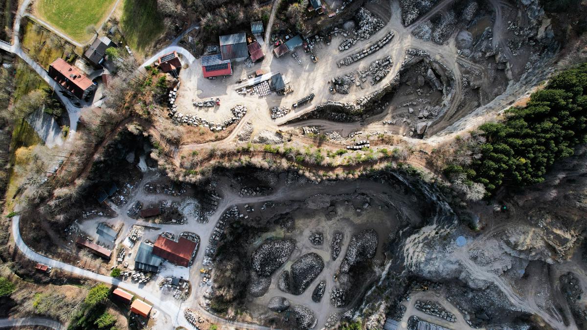 En lodrät flygvy över ett stenbrott: ljus skuren berggrund, staplar av hällar, slingrande grusvägar och några verkstadsbyggnader med röda tak, med skog i kanterna.