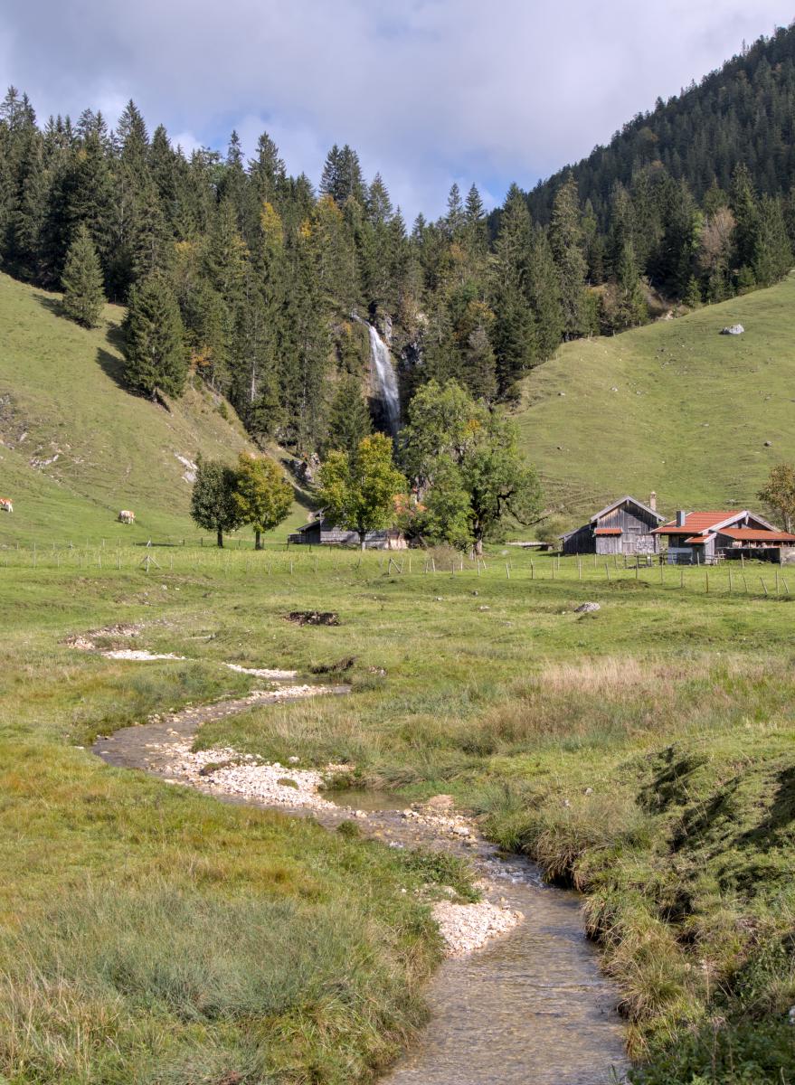 Ein Wasserfall und einige Berghütten befinden sich im Hintergrund. Der Bach des Wasserfalls schlängelt sich zur Kamera hin. Im Hintergrund sind die Berge zu sehen.