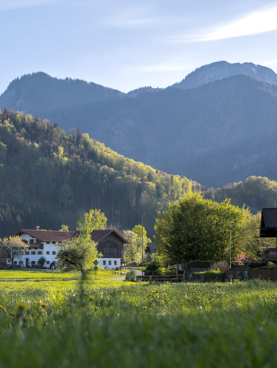 An old farm building next to the Bavarian Alps. The sky is bluish with some white clouds. There is a road snaking through the image.
