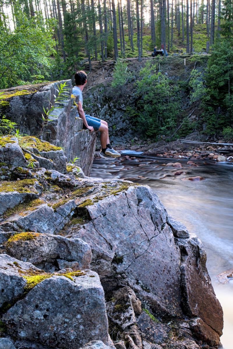 Ein Selbstporträt von mir, wie ich auf einem Felsen neben einem Wasserfall sitze. Der Wasserfall ist wegen der Langzeitbelichtung verwaschen.