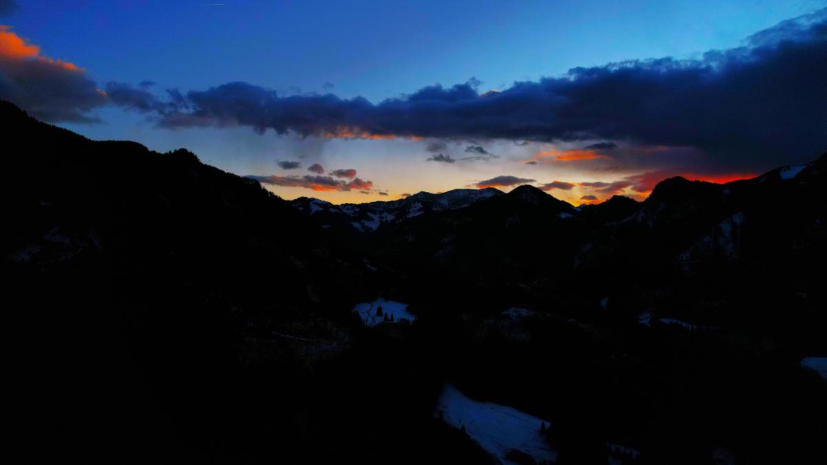 Mörka bergssiluetter ramar in en smal dal i skymningen; ett band av orangebelysta moln glöder ovanför de snöstrimmiga kammarna i fjärran.