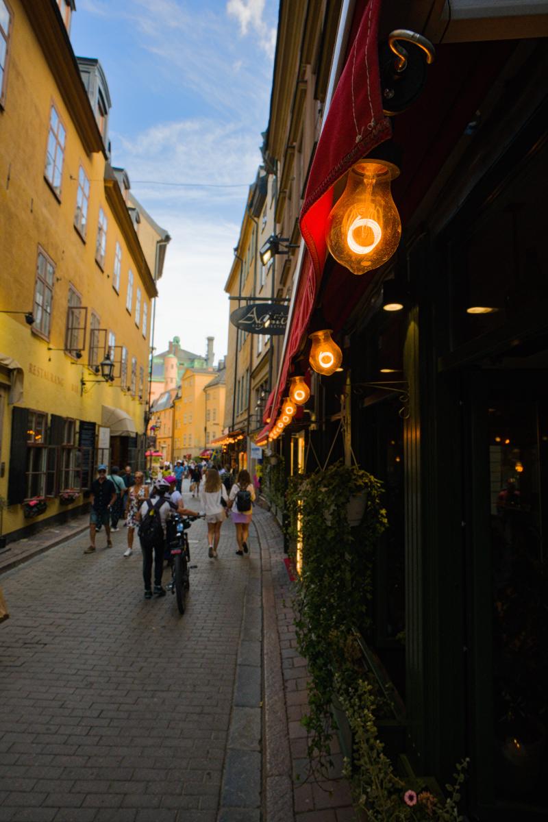 A narrow cobbled lane in Gamla stan, Stockholm's old town, on a summer afternoon. On the right, a row of glowing vintage light bulbs hangs beneath a red awning above a restaurant's dark front with trailing ivy, leading the eye down the street, while yellow and ochre houses line the left side. Pedestrians and a cyclist pushing a bike walk away toward pastel buildings under a blue sky with light clouds.